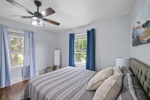 A bedroom with a bed, a ceiling fan, a window with curtains, and a painting on the wall.