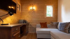 Un acogedor interior de cabaña con una pared de madera, un escritorio con lámpara, una impresora, un sofá con cojines y una ventana.