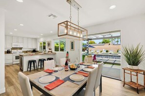 A modern kitchen with white cabinets, a dining area with a wooden table set for six, and a view of an outdoor pool area. - Saltwater Pool & Spa, Fire Pit, Expansive Mountain View (Palm Springs)