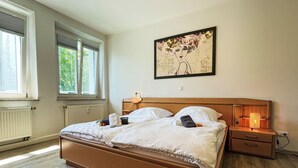 A bedroom with a wooden bed, two bedside tables, a nightstand with a lamp, and a large window with blinds.