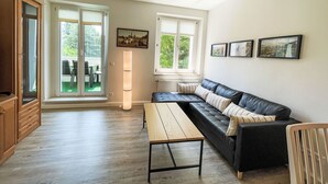 A living room with a wooden floor, a black leather sofa, a wooden coffee table, and a large window with blinds.