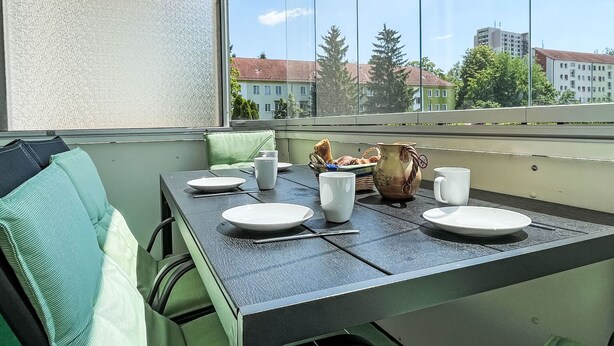 A dining area with a table set for two, a green cushioned chair, and a view of buildings through a window.