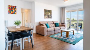 A modern living room with a wooden dining table, a beige sofa with teal cushions, and a coffee table. There are two paintings on the walls and a view of the outdoors through a sliding glass door. - GuestReady - Caniço city view (Caniço)