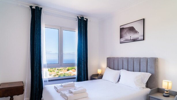 A hotel room with a bed, two bedside tables, a nightstand, a window with curtains, and a framed picture on the wall. - GuestReady - Caniço city view (Caniço)