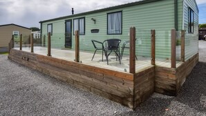 Une caravane verte avec une terrasse entourée d’une clôture, deux chaises et une fenêtre.