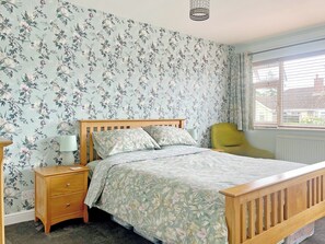 A bedroom with a floral wallpaper, a wooden bed, a nightstand, a lamp, a green chair, and a window with blinds. - 2 bedroom accommodation in Bottesford (Bottesford)