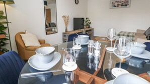 A dining area with a glass table set for a meal, surrounded by chairs. A television is mounted on the wall, and there is a plant on a shelf. - Beachcomb (Weymouth)