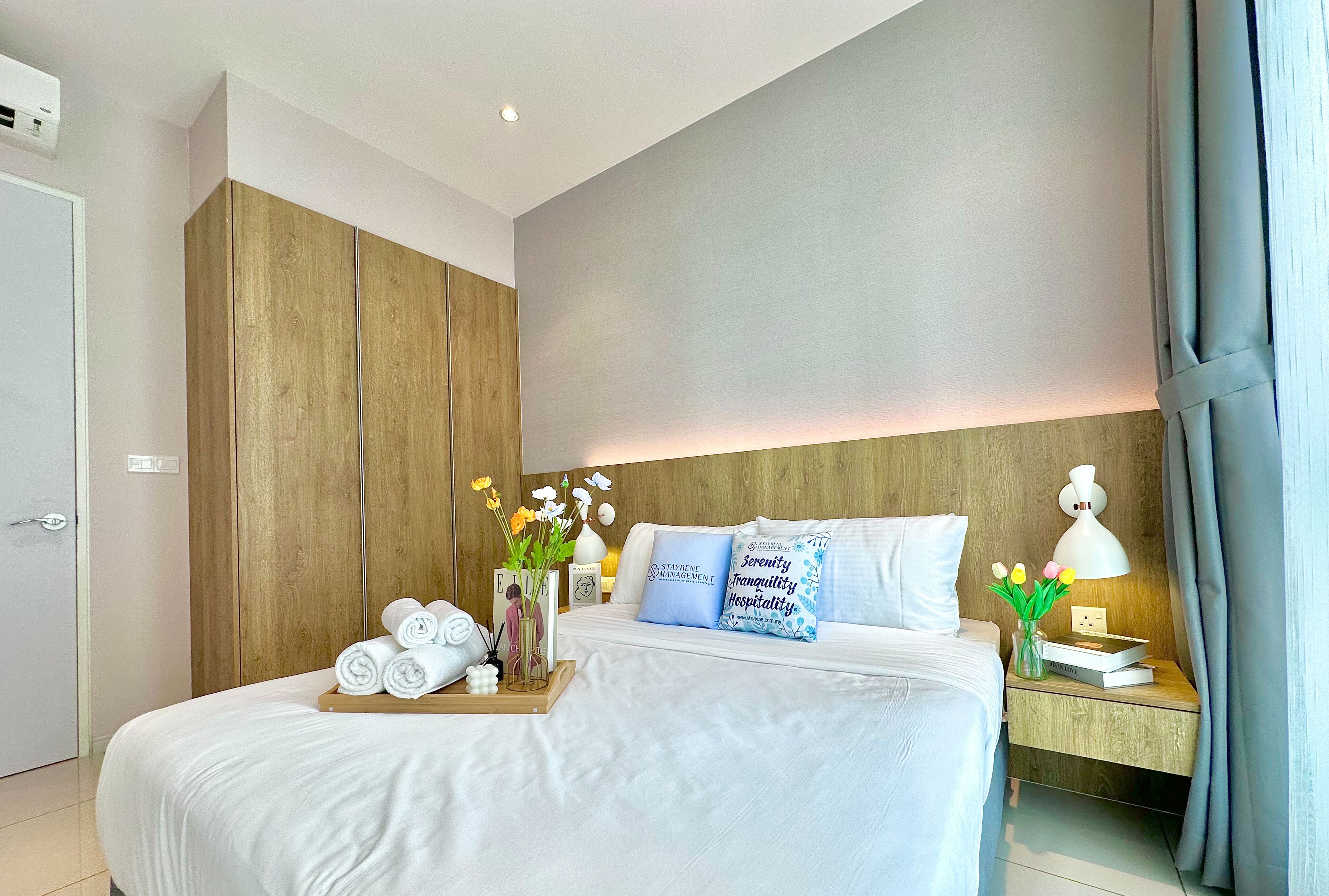A hotel room with a large bed, wooden wardrobe, bedside table with a lamp, and a tray with towels and flowers.