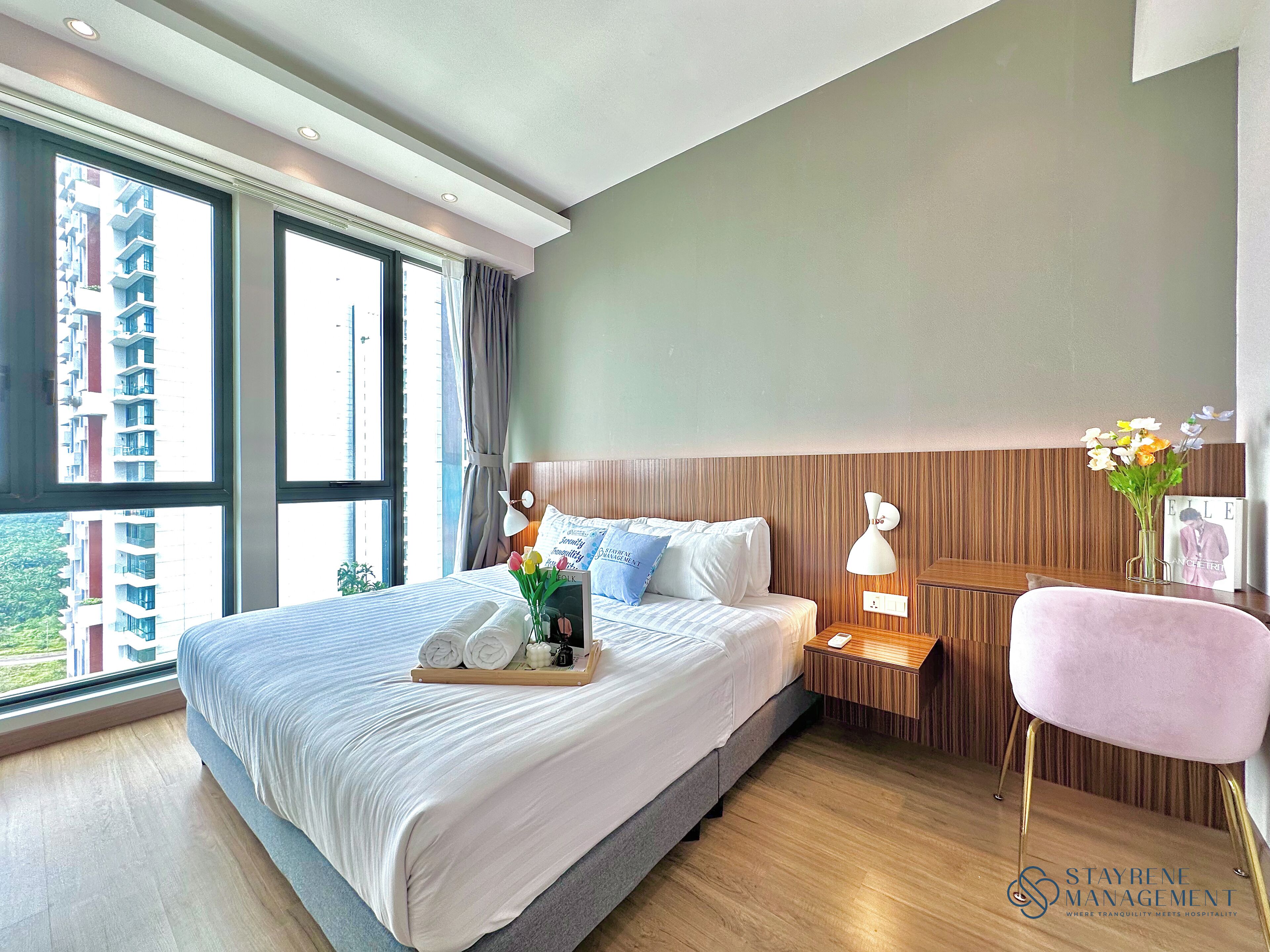 A modern hotel room with a large bed, a bedside table with a lamp, a chair, and a view of the city through large windows.