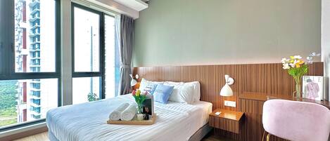 A modern hotel room with a large bed, a bedside table with a lamp, a chair, and a view of the city through large windows.