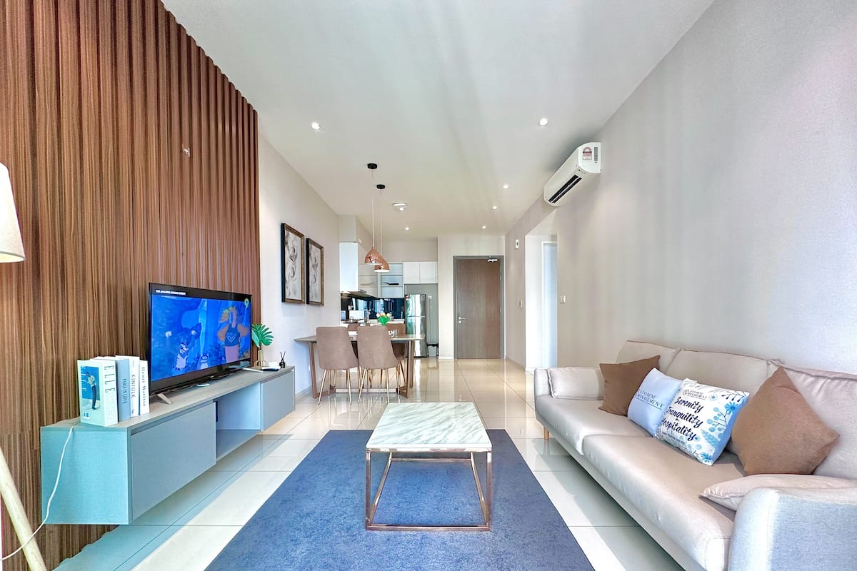 A modern living room with a flat-screen TV, a sofa, a coffee table, and a dining area with chairs and a kitchen in the background.