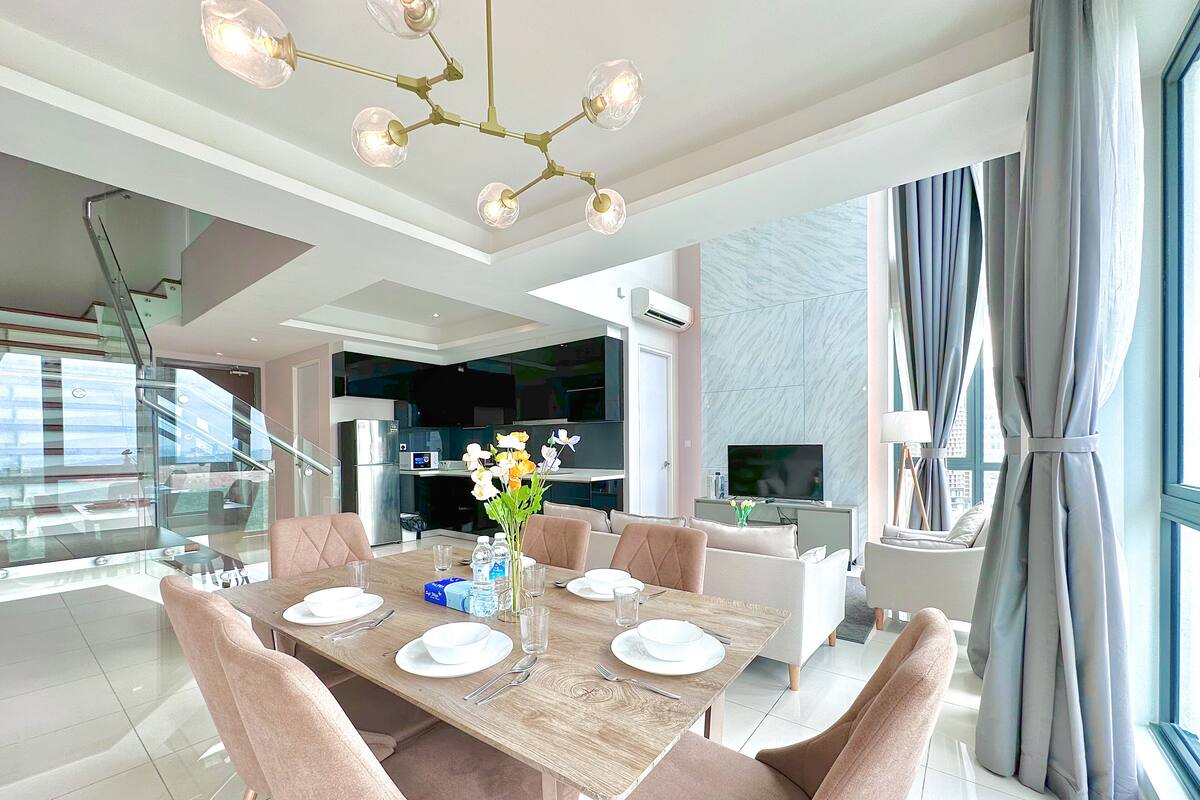 A modern dining area with a wooden table set for six, surrounded by cushioned chairs. A chandelier hangs above, and a vase with flowers is on the table.