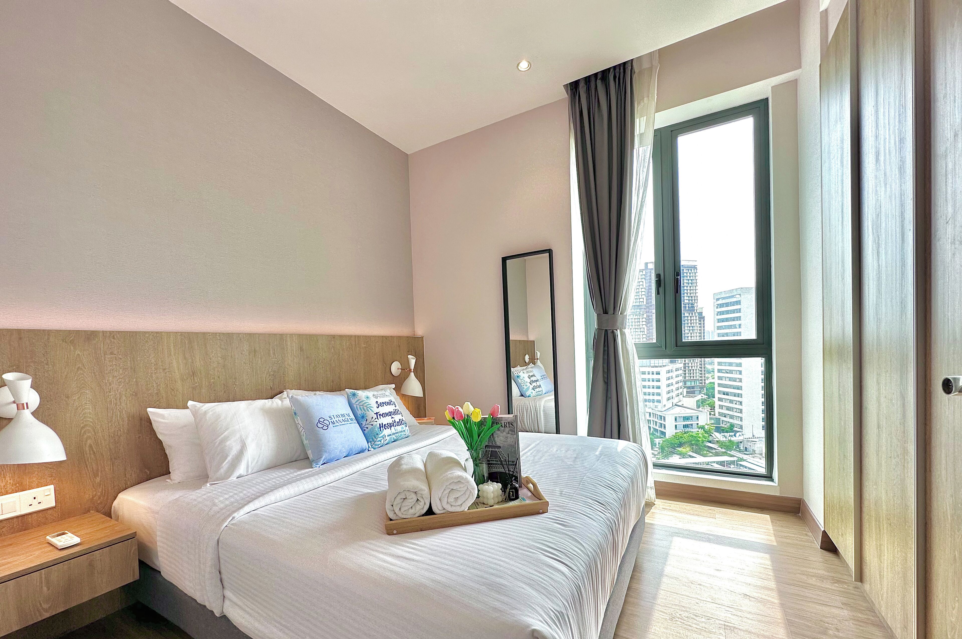 A hotel room with a large bed, a bedside table with a lamp, a tray with towels and toiletries, and a window with a city view.
