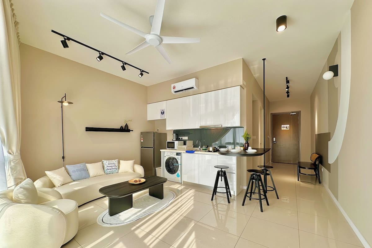 A modern living room with a sofa, coffee table, and a dining area with stools. There is a kitchen with a washing machine and a ceiling fan.