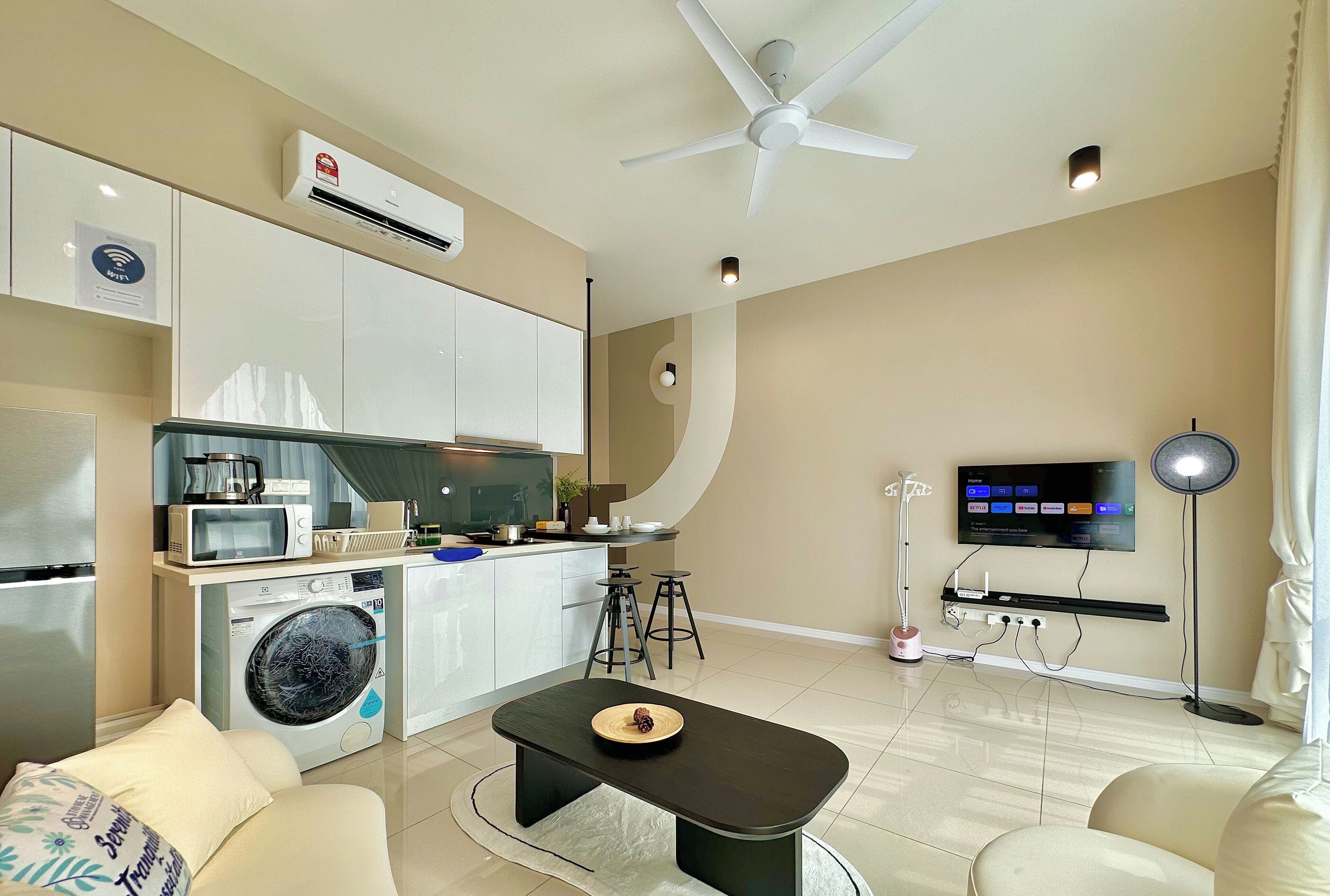 A modern living room with a sofa, a coffee table, a TV, a washing machine, and a kitchen area with a microwave and a kettle.