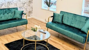 Een woonkamer met twee groene banken, een glazen salontafel, een zwart tapijt, houten vloeren, een marmeren muur en een plafondlamp.