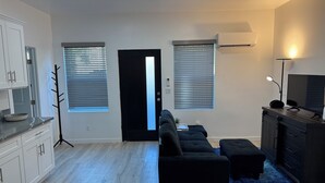 Una sala moderna con un sofá negro, un televisor y una alfombra azul. Hay dos ventanas con persianas, una puerta y una zona de cocina con armarios.