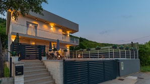 A modern two-story house with a balcony, a gated entrance, and outdoor seating.