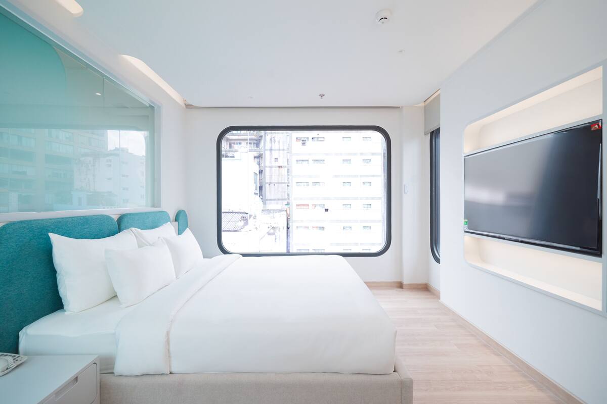 A modern hotel room with a large bed, a television mounted on the wall, and a window offering a city view.