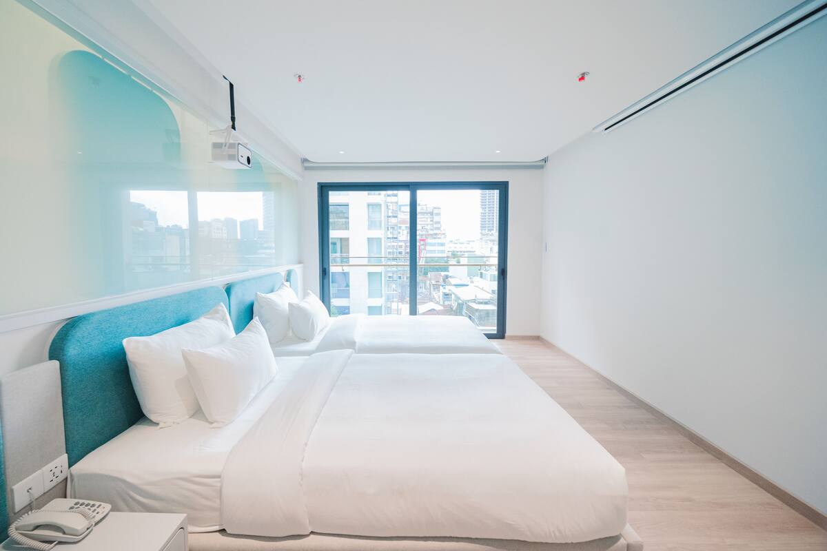 A modern hotel room with a large bed, a bedside telephone, and a city view through a large window.