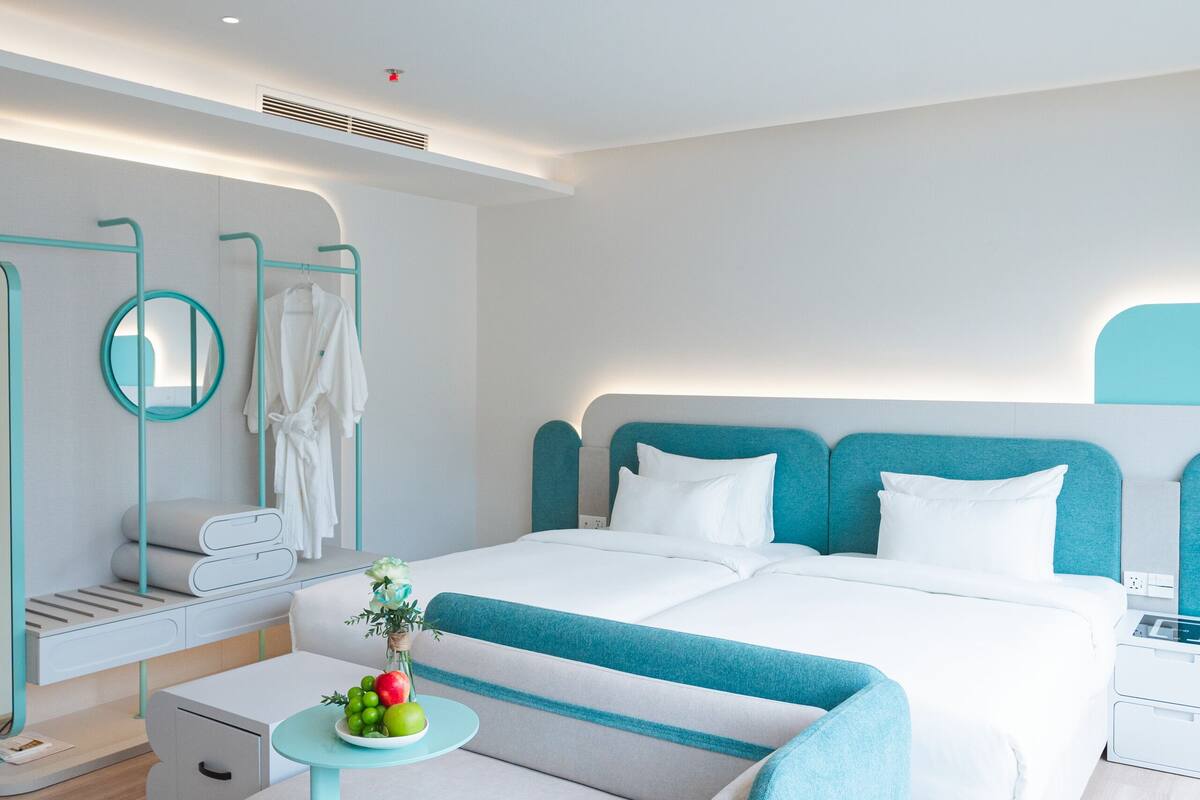 A modern hotel room with a large bed, a small round table with fruit, a desk, and a chair.