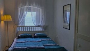 A narrow room with a bed, a window with sheer curtains, a lamp, and a framed picture on the wall.