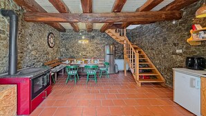 A rustic kitchen with a stone wall, wooden beams, a dining area with green chairs, a wooden staircase, and a red stove.