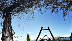 Una terraza de madera con una silla mecedora, una hamaca colgante, y vistas a una piscina y montañas.