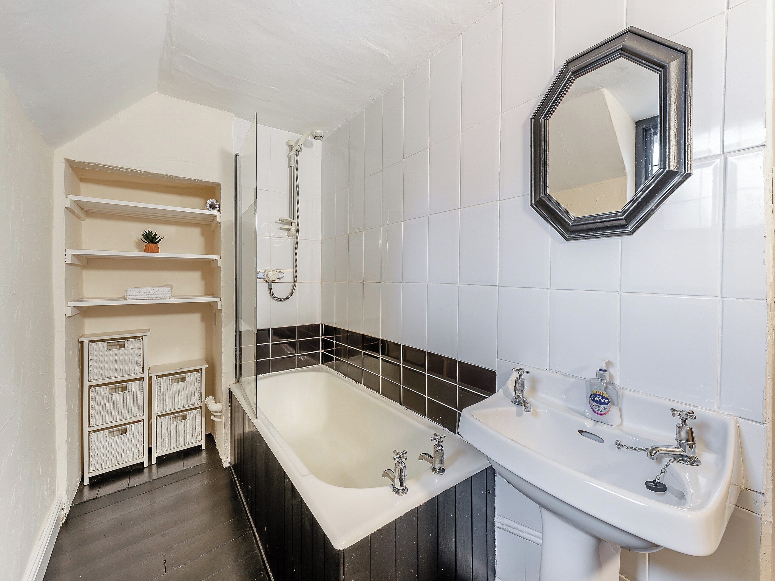 Een badkamer met een bad, een wastafel en een douche. Er zijn planken met manden en een kleine plant.