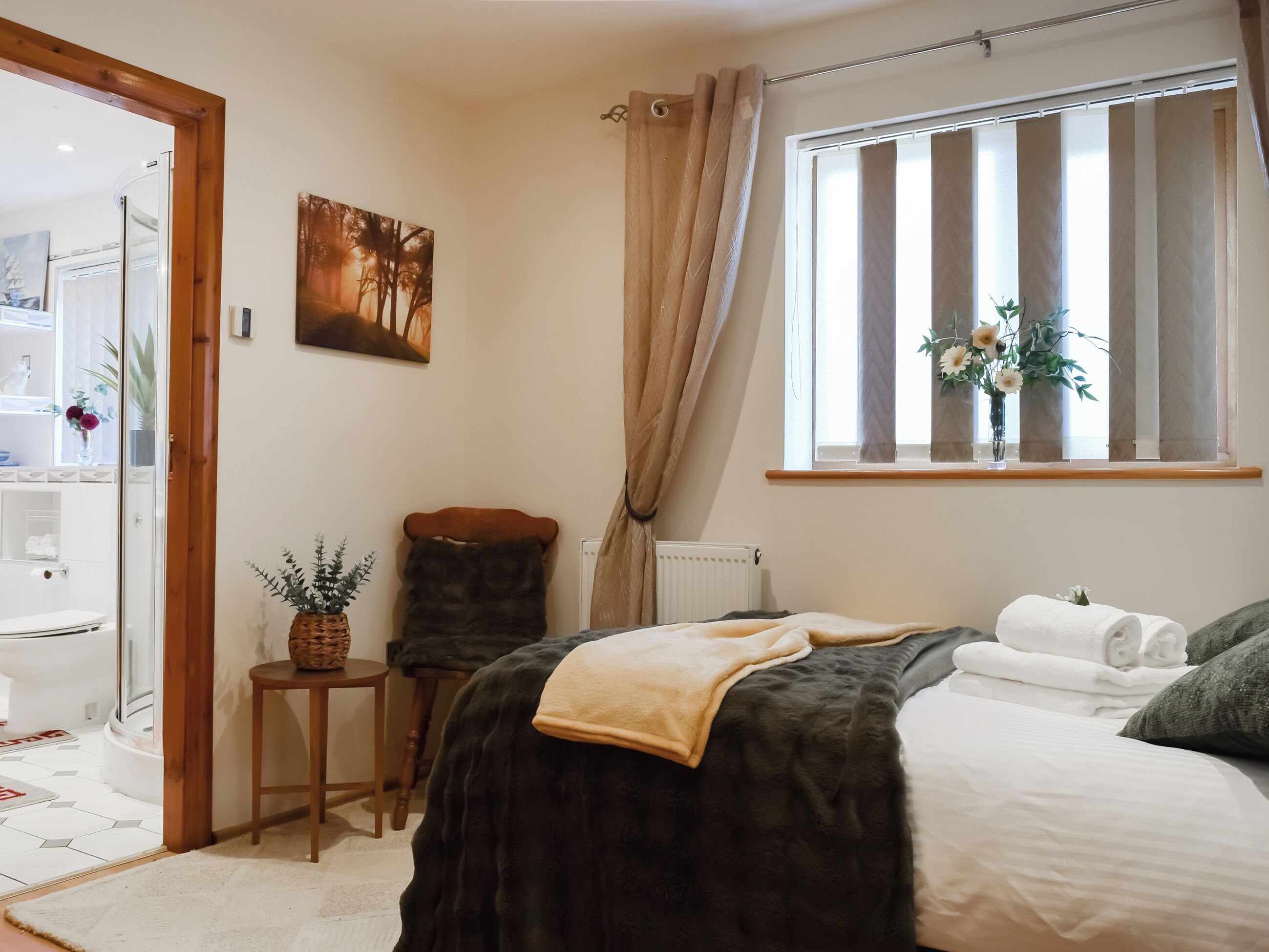 A bedroom with a bed, a window with curtains, a radiator, a small table with a plant, and a painting on the wall.