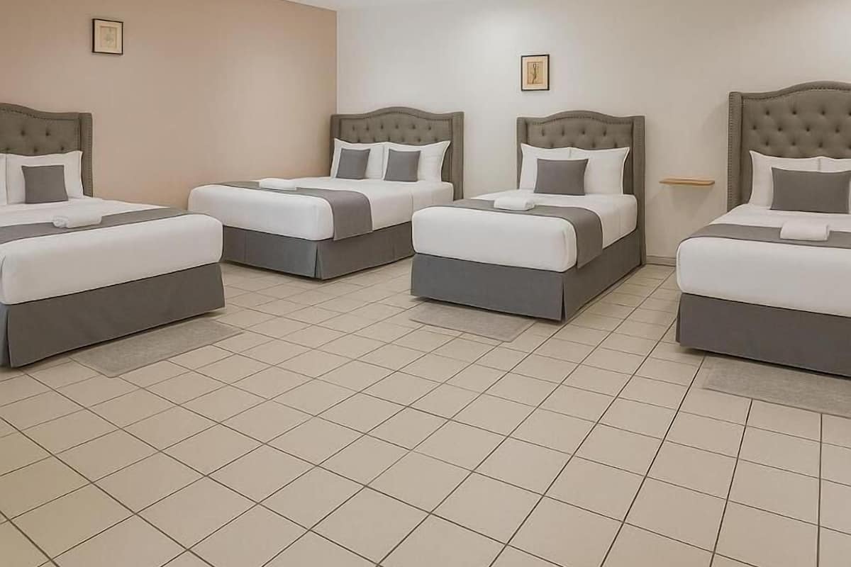 Een hotelkamer met vier bedden op een rij, elk met een grijze hoofdbord en witte beddengoed. De kamer heeft een betegelde vloer en beige wanden.