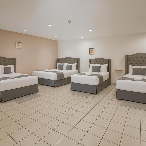 Habitación de hotel con cuatro camas dispuestas en fila, cada una con cabecero gris y ropa de cama blanca. El piso es de baldosas y las paredes son color beige.