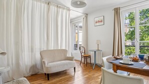 Quarto aconchegante com piso de madeira, poltrona bege, mesa pequena com abajur e vista para o verde lá fora.