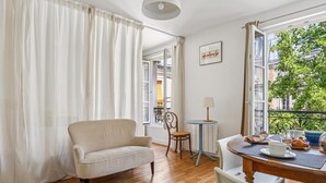 Quarto com sofá, uma mesinha com xícara e doces, e vista para uma varanda com vegetação.