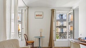 Um quarto acolhedor com um sofá, uma mesinha redonda com abajur, uma cadeira e vista para uma varanda com edifícios.