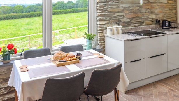Una cocina moderna con una pared de piedra, un comedor con una mesa dispuesta para dos personas, y vistas a un paisaje verde a través de amplios ventanales.