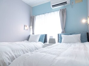 Habitación de hotel con dos camas, una ventana con cortinas, un equipo de aire acondicionado y una mesita de noche con lámpara.