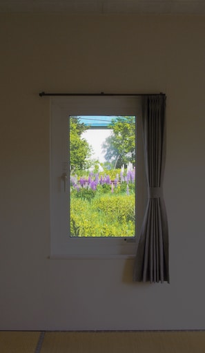 A room with a window, a curtain, and a view of a garden with flowers. - FURANO RETREAT (Furano)