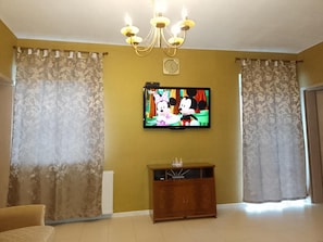 A living room with a television displaying animated characters, a chandelier, curtains, and a wooden cabinet.