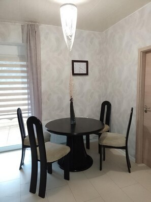 A dining area with a round table, four chairs, a wall clock, and a window with blinds.
