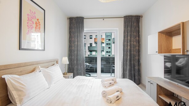 A hotel room with a bed, a TV, a wooden cabinet, and a view of buildings through a glass door. - MoHost - Lube (Brasov)
