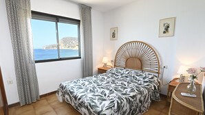 Une chambre à coucher avec un lit, une fenêtre donnant sur la mer, une table de chevet avec une lampe et un objet décoratif fixé au mur.