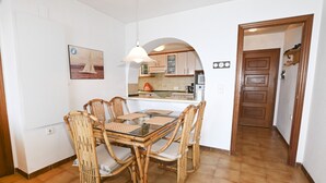 Un coin repas avec une table à plateau de verre et quatre chaises en osier, une cuisine avec des meubles en bois et une porte en bois donnant sur une autre pièce.