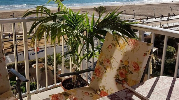 A balcony with a view of the beach, featuring a potted plant, a chair with floral upholstery, and red umbrellas. - Attractive Apartment in El Campello in Beachfront (El Campello)