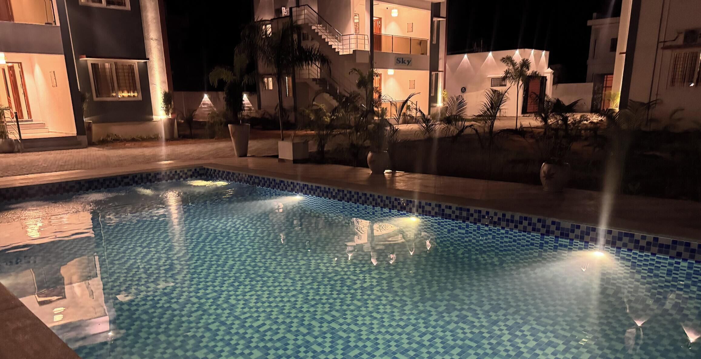 Piscina com padrão de azulejos quadriculados, rodeada por um edifício bem iluminado com varandas e uma placa com a inscrição "Sky".