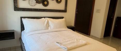 Una cama bien tendida con sábanas blancas, una obra de arte decorativa en la pared encima y una puerta a la derecha.