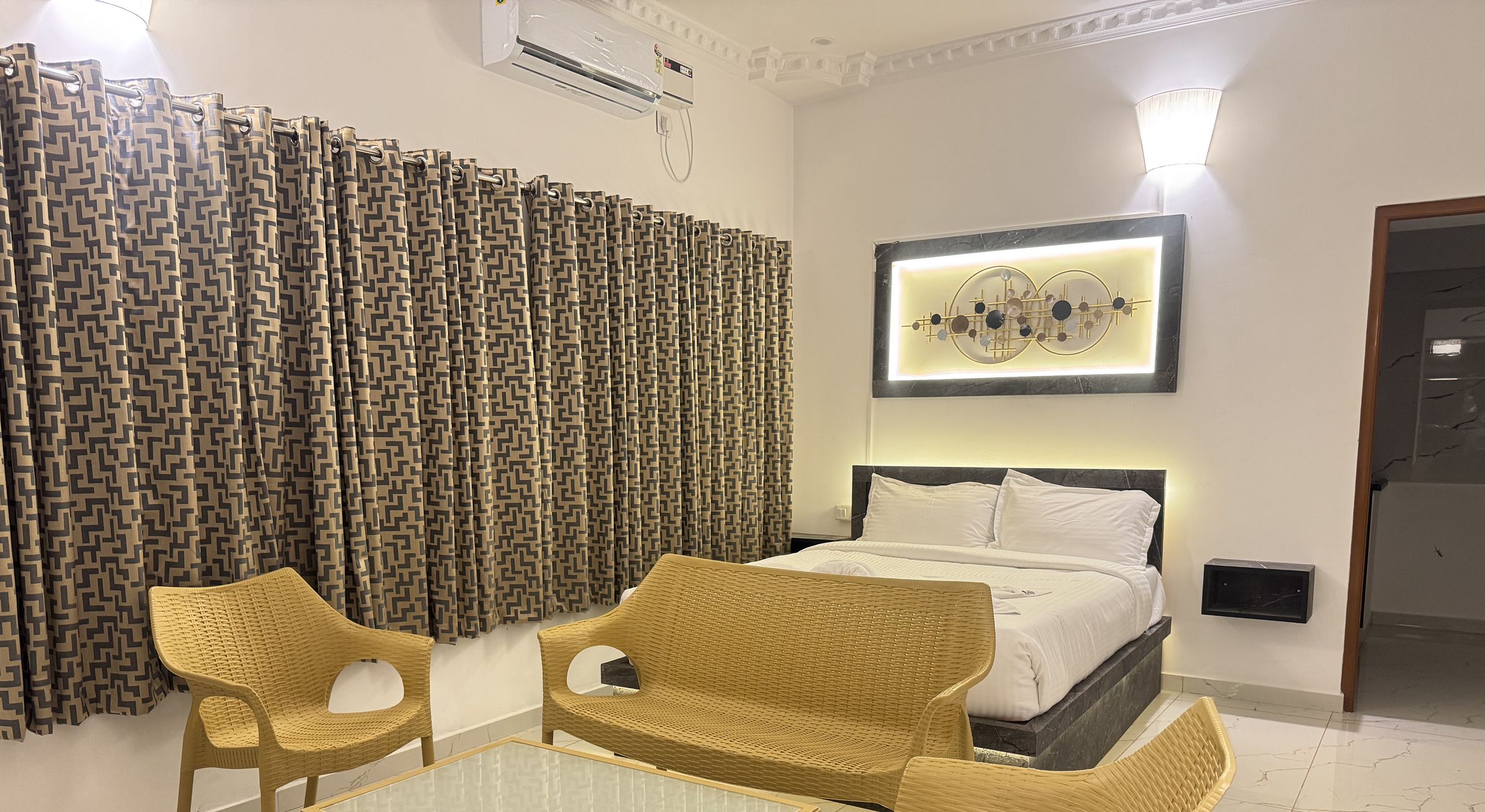 A hotel room with a bed, two wicker chairs, a glass-top table, and a patterned curtain.