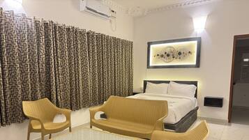 A hotel room with a bed, two wicker chairs, a glass-top table, and a patterned curtain.