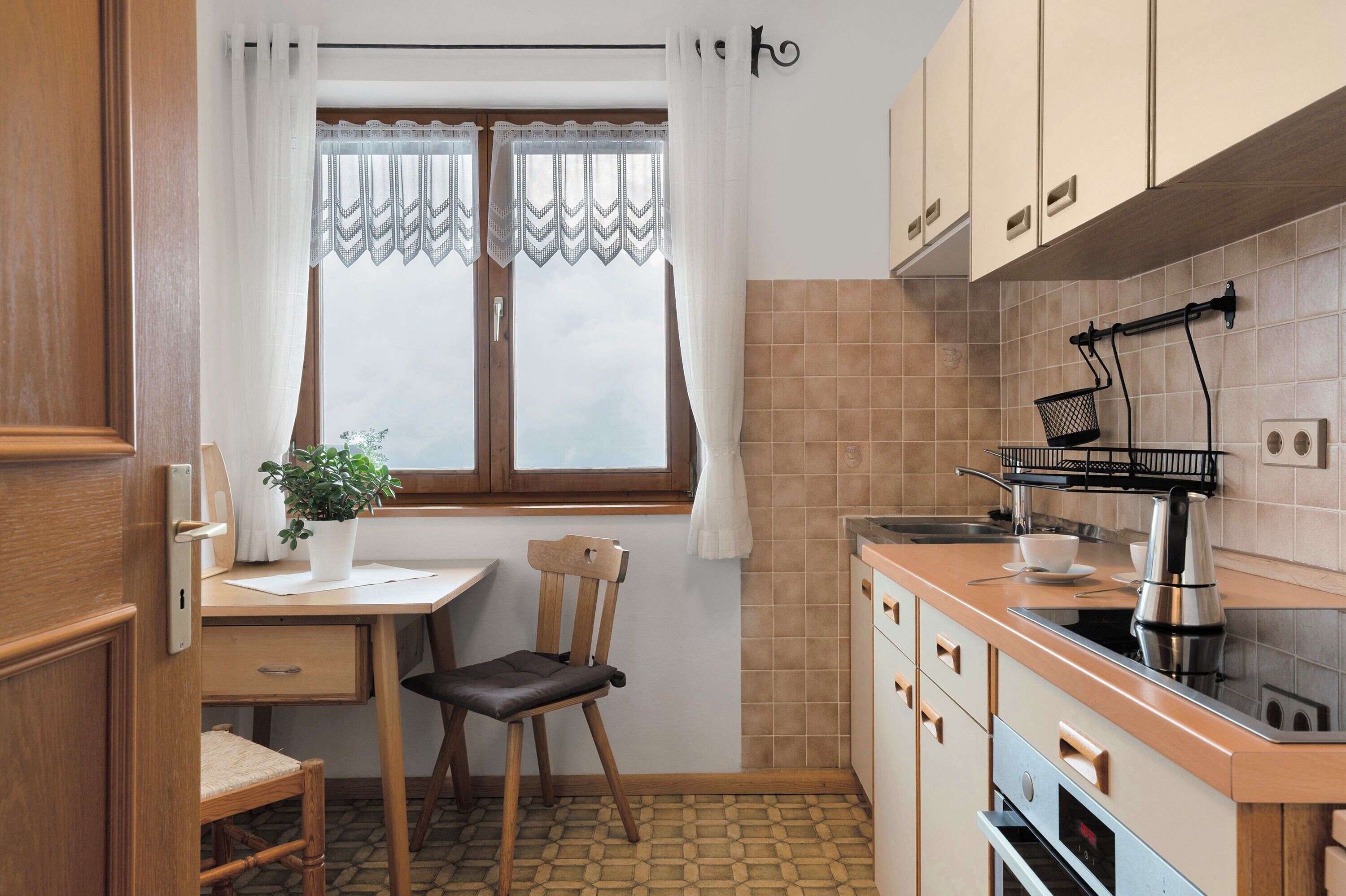 Uma cozinha compacta com mesa de madeira, uma cadeira, uma planta em vaso, janela com cortinas e painel de azulejos.