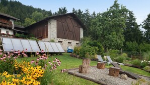 Une grange rustique, dotée d’un toit en pente, entourée d’un jardin bien entretenu où poussent différentes variétés de fleurs, et bordée d’un chemin de gravier.
